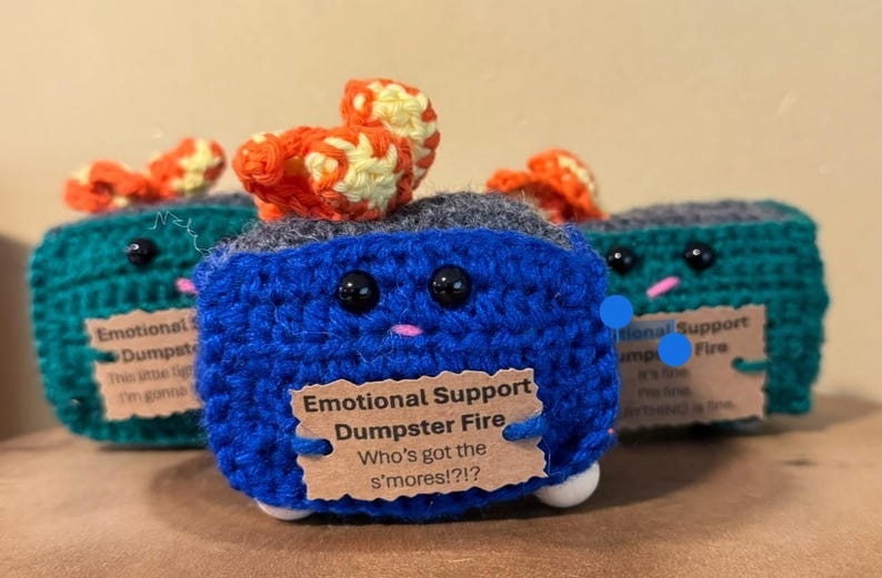 May include: Crocheted blue, green, and gray dumpster fire plush toys with orange and yellow flames. Each has black eyes, a pink mouth, and a tag that reads "Emotional Support Dumpster Fire".