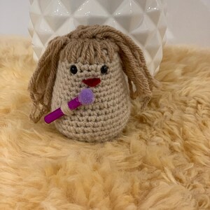 May include: A crocheted doll with brown hair, a purple microphone, and a red mouth. The doll is sitting on a light brown fluffy surface.