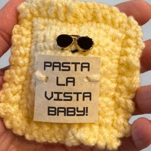 May include: A yellow crocheted ravioli pin, featuring a pair of sunglasses and a tag with the text "PASTA LA VISTA BABY!". This handmade pin is a playful, food-inspired accessory.
