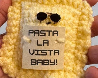 May include: A handmade, yellow crocheted ravioli pin with a pair of sunglasses and a tag that reads "PASTA LA VISTA BABY!". The pin is a fun, food-themed accessory.