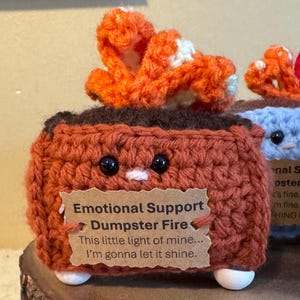 May include: A handmade, crocheted brown dumpster with a smiling face and an orange flame. The dumpster has a tag that reads "Emotional Support Dumpster Fire" and the quote "This little light of mine... I'm gonna let it shine."