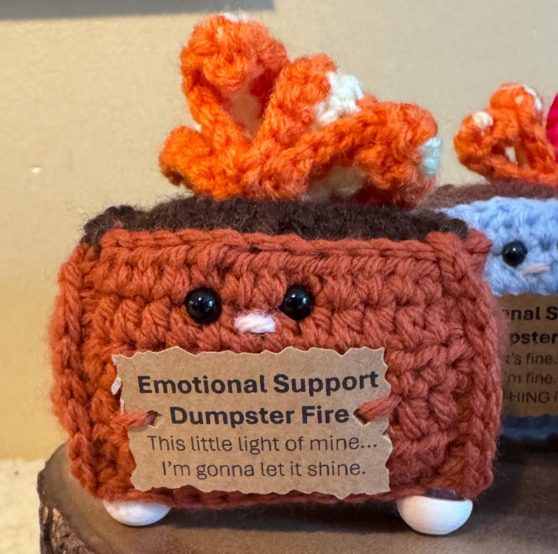 May include: A handmade, crocheted brown dumpster with a smiling face and an orange flame. The dumpster has a tag that reads "Emotional Support Dumpster Fire" and the quote "This little light of mine... I'm gonna let it shine."