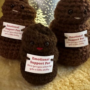 May include: Three brown crocheted poop-shaped figures with smiling faces and small black eyes. Each figure holds a tag that reads "Emotional Support Poo. Here for you when life gets a little shitty."