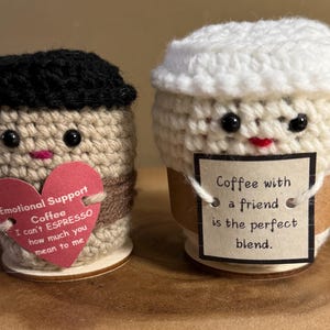 May include: Two crocheted coffee cup characters with faces. One cup is brown with a black crocheted lid and a pink heart with the text "Emotional Support Coffee I can't ESPRESSO how much you mean to me". The other cup is white with a white crocheted lid and a brown paper label with the text "Coffee with a friend is the perfect blend."