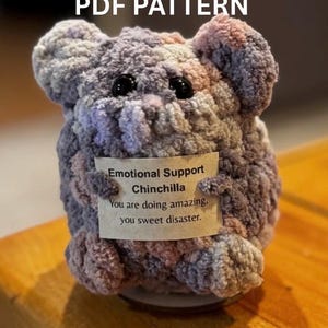 May include: A crocheted chinchilla toy in grey, pink, and purple tones, with black button eyes. It has a tag that says "Emotional Support Chinchilla. You are doing amazing, you sweet disaster." The text "PDF PATTERN" is at the top.