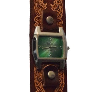Puede incluir: Reloj de cuero marrón con esfera rectangular plateada y esfera verde. La correa del reloj presenta un diseño floral en relieve y un cierre de hebilla. La correa mide aproximadamente 2,5 cm de ancho.