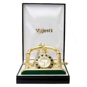 May include: A gold pocket watch with a black face and Roman numeral markings is displayed in a gold stand with a green velvet base. The watch is in a black and white box with the word "MajesTi" printed on the inside.