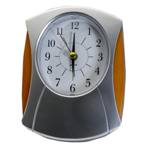 Pode incluir: Um despertador de prata e madeira com um mostrador branco e ponteiros pretos. O relógio está ajustado para 12:00.