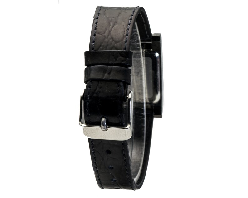 Peut inclure: Une montre-bracelet noire avec un cadran rectangulaire et un bracelet en cuir noir. Le bracelet pr&eacute;sente un motif textur&eacute; et une boucle argent&eacute;e. La montre est pr&eacute;sent&eacute;e sur fond blanc.