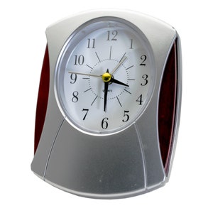 Pode incluir: Um despertador prateado e vermelho com um mostrador branco e ponteiros pretos. O mostrador do relógio tem números pretos e a palavra "Quartz" impressa nele.