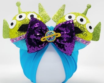 Puede incluir: Diadema turquesa con un lazo de lentejuelas moradas y dos orejas de extraterrestre verdes con ojos negros y pupilas blancas. Las orejas tienen un acabado brillante y un ribete plateado y azul.