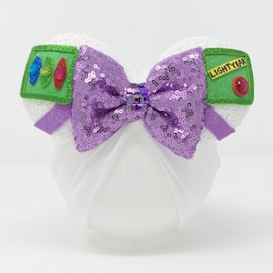 Peut inclure: Oreilles de souris Minnie blanches avec un nœud en sequins violets et un patch vert qui dit "LIGHTYEAR" avec un bouton rouge.