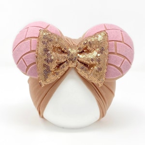 May include: A brown turban headband with a large gold sequin bow and two pink felt ears shaped like a Mexican sweet bread, concha.