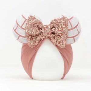 Op de afbeelding: Een blush roze hoofdband met een pailletten strik en twee witte en roze volleybal-thema oren. De hoofdband is om een witte vorm gewikkeld, waardoor een speels en sportief accessoire ontstaat.