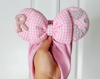 May include: Pink headband with a large bow featuring a pink and white gingham pattern. The bow is adorned with a rhinestone-studded letter "R", a heart, a Mickey Mouse head, and other decorative elements.