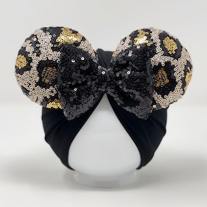 May include: Black turban with a large black bow and two leopard print sequined ears. The ears are gold, black, and white.