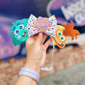May include: A colorful headband featuring characters from the movie Inside Out. The headband has a polka-dot bow with the text "Core Memory Day". The characters include a teal character with a smiling face and an orange character with a wide grin.
