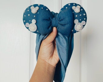 May include: A blue fabric headband with a large bow and two mouse ears decorated with white pearl-like beads and silver glitter Mickey Mouse silhouettes.