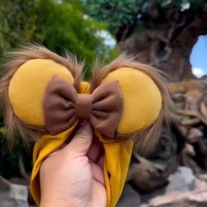 May include: Yellow and brown mouse ear headband with a brown bow. The ears are yellow with brown faux fur trim. The headband is yellow. The accessory is held up in front of a blurred background of trees and sky.