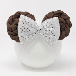 May include: A white turban headband with a large silver sequin bow and two brown braided buns on top.