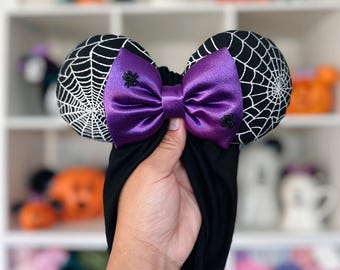 以下が含まれることがあります： 紫と黒のヘッドバンドで、クモの巣のデザインと大きな紫のリボンが付いています。ヘッドバンドは小さな黒いクモで飾られています。
