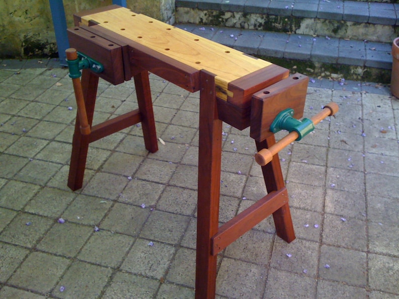May include: A wooden workbench with two vises, one on each end. The workbench is made of dark wood and has a light wood top. The workbench is sitting on a brick patio.