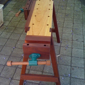 May include: A wooden workbench with a vise and a wooden top. The workbench is made of dark wood and has a light wood top. The vise is made of metal and has a green handle.
