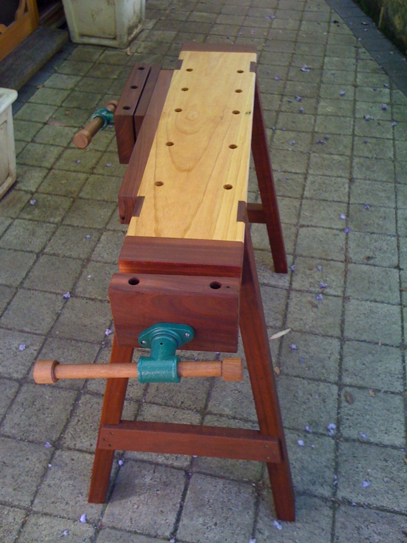 May include: A wooden workbench with a vise and a wooden top. The workbench is made of dark wood and has a light wood top. The vise is made of metal and has a green handle.