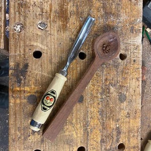 May include: A woodworking scene featuring a chisel with a light-coloured handle and a cherry logo, alongside a partially carved wooden spoon. The spoon is made of dark wood. The tools rest on a well-worn wooden workbench, showcasing the craft.