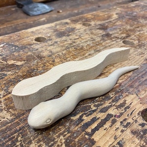 May include: Two wooden snake carvings, one complete and the other a block of wood. The finished snake is light in colour, with a detailed head and body. The unfinished piece is a light-coloured block.