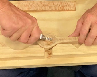 以下が含まれることがあります： 人が小さな彫刻道具で木のスプーンを彫っています。スプーンは木製の型の中に置かれています。スプーンの周りに木くずが見えます。