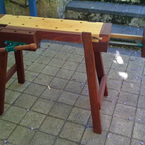 May include: A wooden workbench with a vise and a wooden top. The workbench is made of dark wood and has a light wood top. The workbench is sitting on a patio.