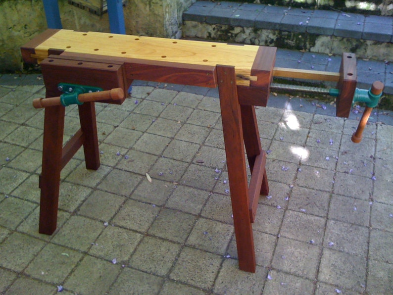 May include: A wooden workbench with a vise and a wooden top. The workbench is made of dark wood and has a light wood top. The workbench is sitting on a patio.