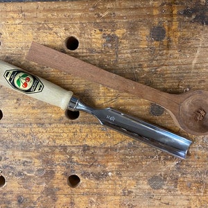 May include: A wooden spoon and a wood carving chisel on a wooden workbench. The spoon is made of dark wood and has a rounded bowl. The chisel has a metal blade and a wooden handle with a cherry logo.