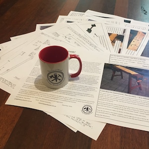 May include: A red and white mug with a logo that says "The Joy of Wood" and a crossed axe design. The mug is sitting on a table with a stack of papers. The papers are blueprints for a woodworking project. The text on the papers says "The Low Stove on Stands / Raw Stand on Stands".