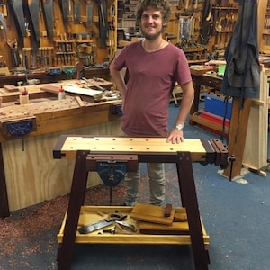 May include: A wooden workbench with a vise and a shelf underneath. The workbench is made of light and dark wood and has a natural finish. The workbench is standing on a blue floor.
