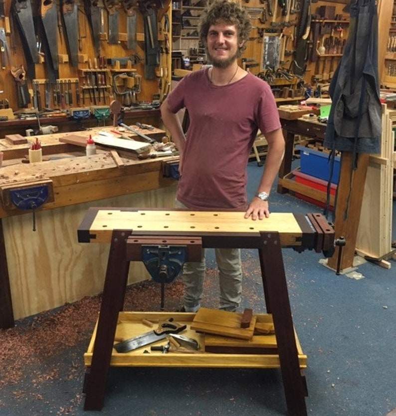 May include: A wooden workbench with a vise and a shelf underneath. The workbench is made of light and dark wood and has a natural finish. The workbench is standing on a blue floor.