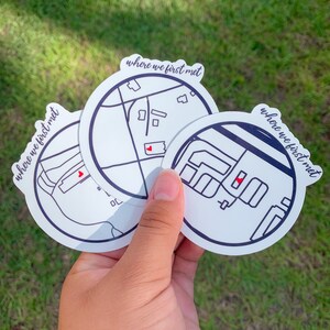 May include: Three white circular stickers with black outlines and the text "where we first met". Each sticker features a map design with a red heart marking a location. The stickers are held against a green background.
