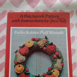 May include: A red book cover with the title "Della Robbia Puff Wreath" features a colorful, handmade wreath with fabric fruit. The wreath is approximately 18 inches (45.7 cm) round. The text "A Patchwork Pattern with Instructions by Yours Truly" is at the top.