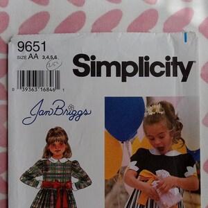 May include: Simplicity sewing pattern number 9651 for a girl's dress. The pattern includes two dress styles, one with a plaid print and the other with black and white stripes. The pattern is for sizes AA, 3, 4, 5, and 6.