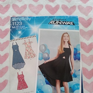 May include: Simplicity sewing pattern number 1123 for a girls' dress. The pattern includes three different styles: a high-low dress with a cutout, a high-low dress with a floral print, and a shorter dress with a floral print. The pattern is designed by Project Runway.
