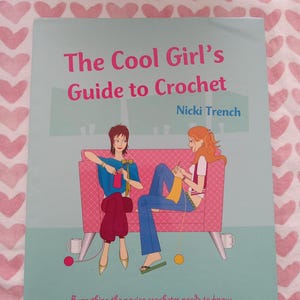 May include: A book cover with the title "The Cool Girl's Guide to Crochet" by Nicki Trench. The cover features illustrations of two women crocheting on a pink couch. The text "Everything the novice crocheter needs to know" is at the bottom.