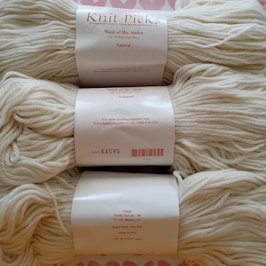May include: Three skeins of natural-colored Knit Picks Wool of the Andes yarn. Each skein has a label with the brand name, yarn type, and care instructions. The yarn is 100% Peruvian wool.