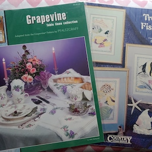 May include: Several cross-stitch pattern books are displayed. One features a table linen collection with a grapevine design. Another showcases a tropical fish trio. The books are surrounded by a table setting with matching grape-themed china.