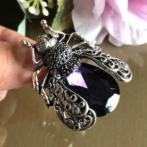 May include: A silver tone brooch in the shape of a bee with a large, faceted, purple crystal. The bee's body is covered in small black rhinestones.