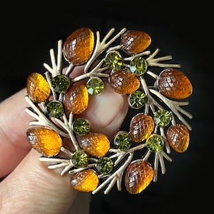 Puede incluir: Un broche circular con un marco metálico en forma de ramas, adornado con piedras de vidrio ovaladas y facetadas de color ámbar y pequeños brillantes redondos de color verde lima dispuestos en forma de corona.