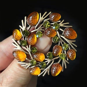May include: A circular brooch featuring a metallic branch-like frame adorned with amber-coloured faceted glass stones shaped like acorns and small, round, bright green rhinestones arranged in a wreath pattern against a dark background.