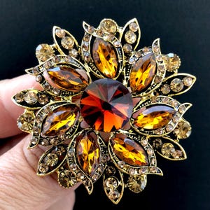 May include: A circular brooch featuring a large, round, deep amber-colored central stone surrounded by multiple marquise-cut amber crystals and smaller round champagne-colored rhinestones set in a gold-toned metal frame with a floral design.