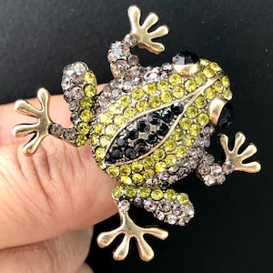Op de afbeelding: Een ring in de vorm van een kikker met een goudkleurige metalen afwerking, bezet met talloze kleine, fonkelende strass-steentjes in limoengroen, antracietgrijs en zwart. De kikker heeft een gedetailleerd lichaam met gespreide poten.