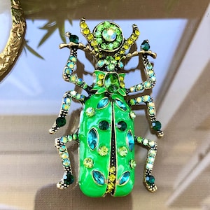 Pode incluir: Um broche em forma de besouro com um corpo de esmalte verde brilhante decorado com strass em vários tons de verde e amarelo. O design do inseto inclui seis pernas, antenas e uma pedra verde oval central na parte superior das costas.